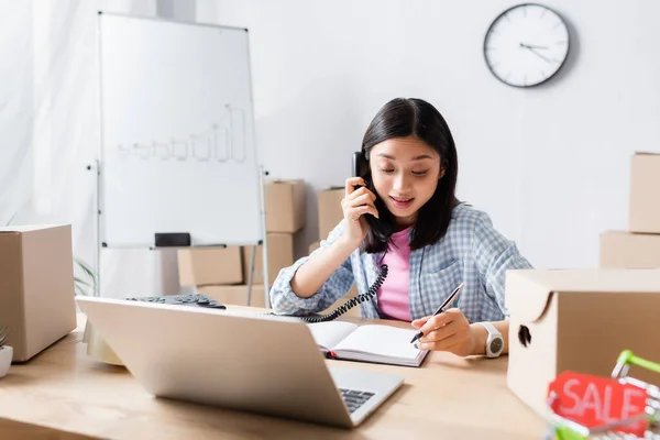 Positive asian volunteer talking on telephone near laptop and carton boxes in charity center on blurred foreground — Stock Photo