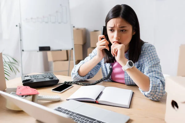 Assustado asiático voluntário falando no telefone e olhando para laptop enquanto sentado na mesa no centro de caridade em primeiro plano borrado — Fotografia de Stock