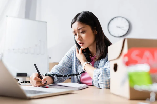 Worried asian volunteer writing in notebook while talking on telephone at desk near laptop on blurred foreground — Stock Photo