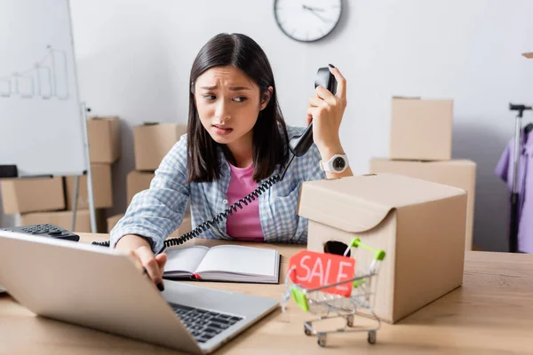 Escéptico asiático voluntario con teléfono mirando caja de cartón cerca de etiqueta de precio con la venta de letras en el carrito de la compra sobre fondo borroso - foto de stock