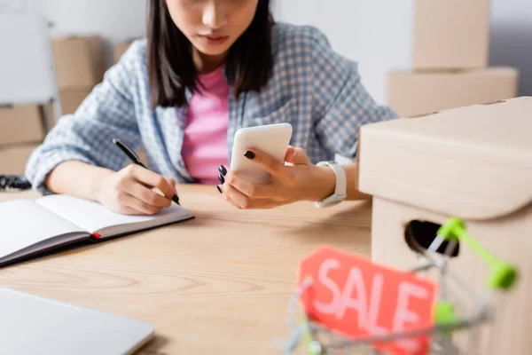 Cropped view of female volunteer with smartphone writing in notebook at desk with blurred shopping cart on foreground — Stock Photo