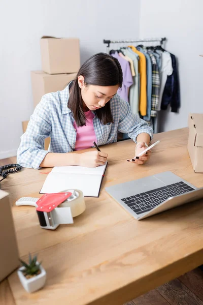 Asian volunteer with smartphone writing in notebook at desk with laptop, carton boxes and scotch dispenser on blurred background — Stock Photo