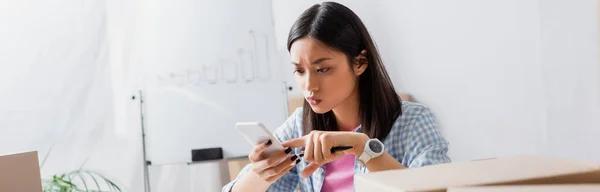 Thoughtful asian volunteer looking at smartphone in charity center on blurred background, banner — Stock Photo