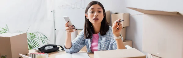 Bénévole asiatique avec smartphone pointant avec stylo à boîte en carton dans le centre de charité, bannière — Photo de stock