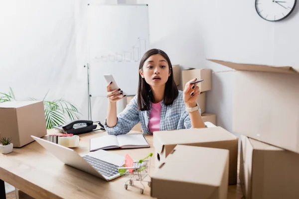 Asian volunteer with smartphone pointing with pen at carton box while sitting at desk with devices in charity center — Stock Photo