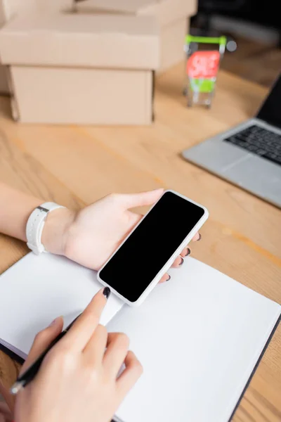 Vista ritagliata della donna che tiene smartphone vicino al notebook alla scrivania con scatole di cartone su sfondo sfocato — Foto stock