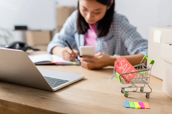 Price tag with sale lettering in shopping cart near carton boxes and laptop with blurred asian woman on background — Stock Photo