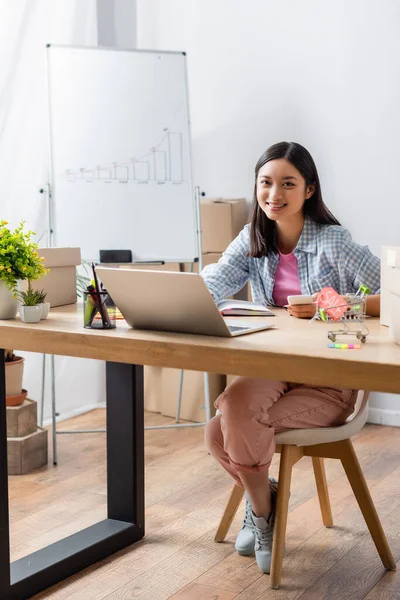 Pleine longueur de sourire asiatique bénévole avec smartphone regardant la caméra tout en étant assis au bureau avec ordinateur portable dans le centre de charité — Photo de stock