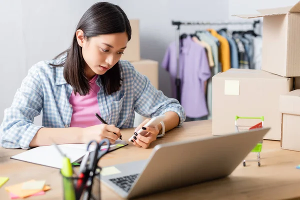 Asian volunteer writing in notebook while looking at smartphone at desk with laptop and carton boxes on blurred foreground — Stock Photo