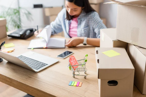 Étiquette de prix avec le lettrage de vente dans le panier près des boîtes de carton sur le bureau avec le bénévole asiatique flou sur le fond — Photo de stock