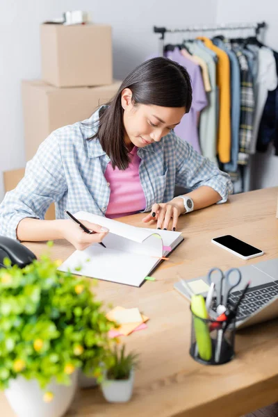 Smiling asian volunteer leafing through notebook while sitting at desk with devices in charity center on blurred foreground — Stock Photo