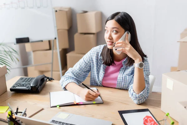 Positiver asiatischer Freiwilliger mit Stift schaut weg, während er am Schreibtisch mit Notizbuch, Geräten und Kartons auf dem Smartphone spricht — Stockfoto