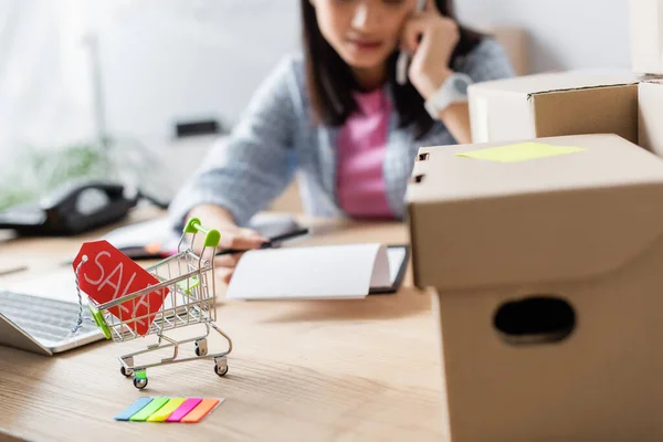 Close up view of price tag with sale lettering in shopping cart near carton boxes on desk with blurred woman on background — Stock Photo