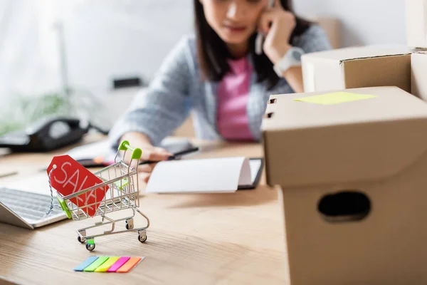 Nahaufnahme von Preisschild mit Verkauf Schriftzug im Warenkorb in der Nähe von Kartons auf dem Schreibtisch mit verschwommener Frau auf dem Hintergrund — Stockfoto