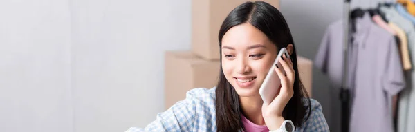 Sonriente asiático voluntario hablando en el teléfono inteligente en el centro de caridad sobre fondo borroso, bandera - foto de stock