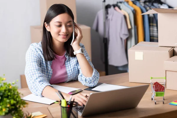 Sorridente volontario asiatico parlando su smartphone mentre si utilizza il computer portatile alla scrivania vicino scatole di cartone con centro di beneficenza offuscata su sfondo — Foto stock