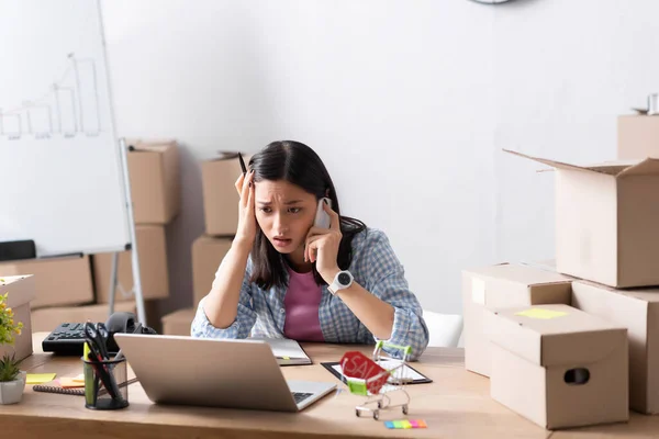 Worried asian volunteer talking on smartphone while looking at laptop at desk with stationery and carton boxes in charity center — Stock Photo