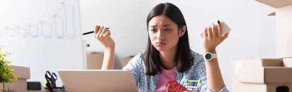 Disappointed asian volunteer with pen and smartphone, looking at laptop near carton boxes in charity center, banner — Stock Photo