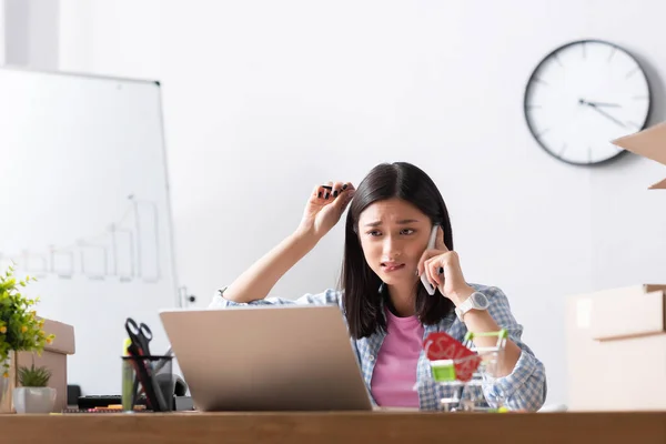 Scared asian volunteer talking on smartphone while looking at laptop near carton boxes in charity center on blurred foreground — Stock Photo