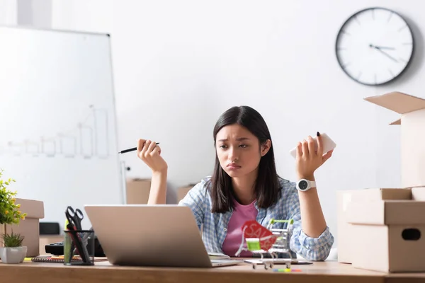 Disappointed asian volunteer looking at laptop, while sitting at desk near carton boxes on blurred foreground — Stock Photo