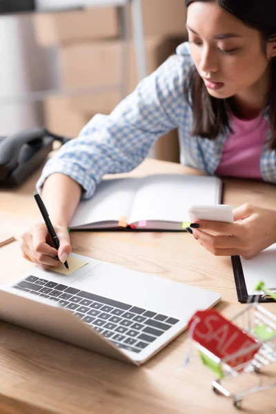 Asiatischer Freiwilliger mit Smartphone-Schriftzug auf Aufkleber, während er mit Notizbuch auf verschwommenem Hintergrund am Schreibtisch sitzt — Stockfoto