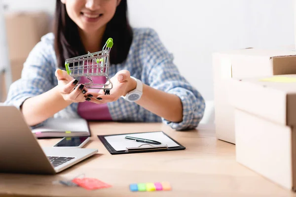 Vista recortada de la mujer que muestra el carrito de compras mientras está sentado en el escritorio con dispositivos y cajas de cartón en primer plano borroso - foto de stock