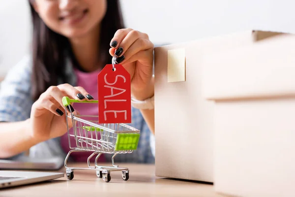 Close up view of price tag with sale lettering in hands of woman near shopping cart and carton boxes on desk with blurred background — Stock Photo