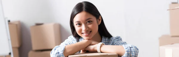 Sonriente asiático voluntario mirando la cámara mientras se apoya en caja de cartón en el centro de caridad sobre fondo borroso, pancarta - foto de stock