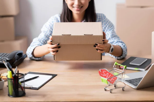 Ausgeschnittene Ansicht einer Frau, die einen Karton hält, während sie am Schreibtisch sitzt, mit Preisschild im Einkaufswagen auf verschwommenem Hintergrund — Stockfoto