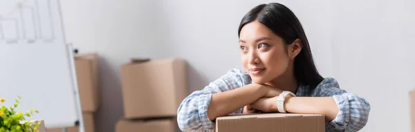 Positive asian volunteer looking away while leaning on carton box in charity center on blurred background, banner — Stock Photo