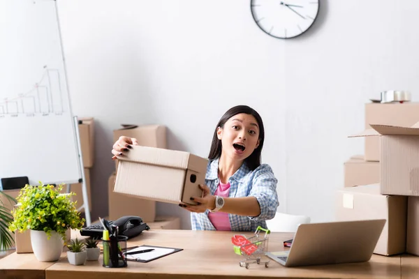 Excited asian volunteer with open mouth showing carton box at desk in charity center — Stock Photo