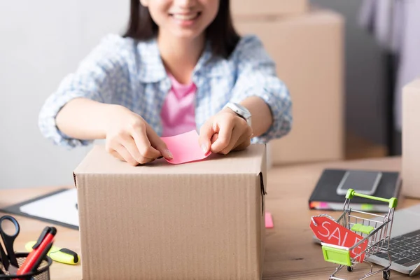 Cropped view of volunteer pasting sticker on carton box near shopping cart while sitting at desk on blurred background — Stock Photo