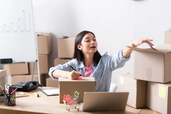 Asian female volunteer with pen, touching carton cardboard while sitting at desk with stationery and devices in charity center — Stock Photo