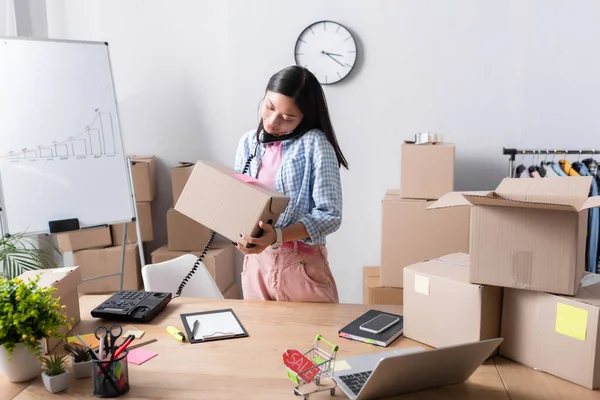 Positivo asiatico volontario con pacchetto di parlare al telefono mentre in piedi vicino scatole di cartone in centro di beneficenza — Foto stock