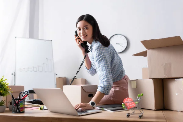 Lächelnder asiatischer Freiwilliger blickt in die Kamera und telefoniert, während er auf dem Schreibtisch neben Kartons im Charity Center sitzt — Stockfoto