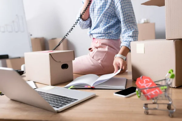 Cropped view of woman talking on telephone and leafing through notebook while sitting near carton boxes on blurred background — Stock Photo