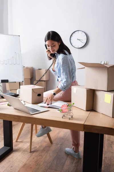 Full length of positive asian volunteer talking on telephone and looking at notebook while sitting near carton boxes in charity center — Stock Photo