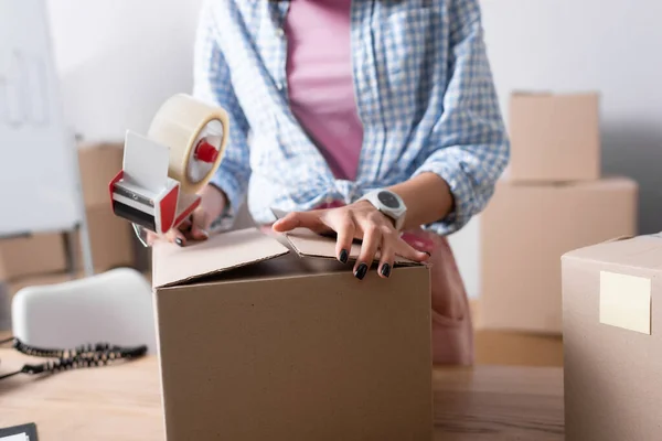 Vue recadrée du bénévole féminin avec distributeur de scotch fermeture boîte en carton dans le centre de bienfaisance sur fond flou — Photo de stock