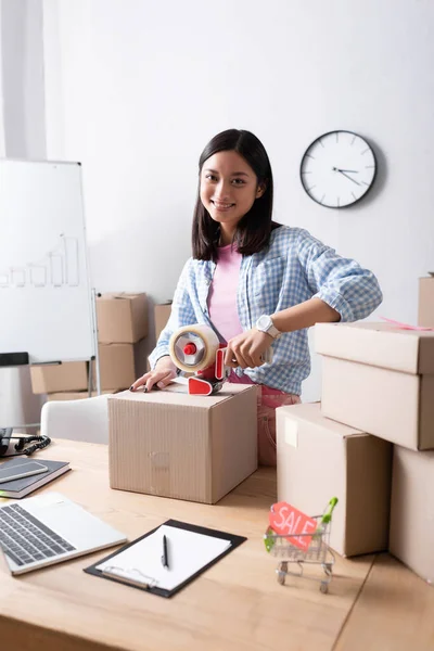 Smiling asian volunteer with scotch dispenser looking at camera while closing package near carton boxes in charity center — Stock Photo