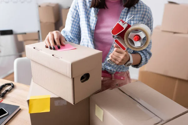 Cropped view of female volunteer holding scotch dispenser near carton boxes in charity center on blurred background — Stock Photo