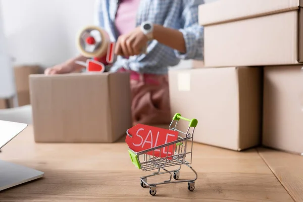 Close up view of price tag with sale lettering in shopping cart on desk near carton boxes with blurred woman on background — Stock Photo