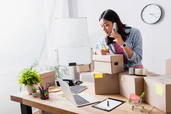 Asian volunteer talking on smartphone near packages, laptop and clipboard on table — Stock Photo