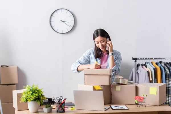 Smiling asian volunteer writing on box near laptop, toy shopping cart and price tag with sale lettering in charity office — Stock Photo