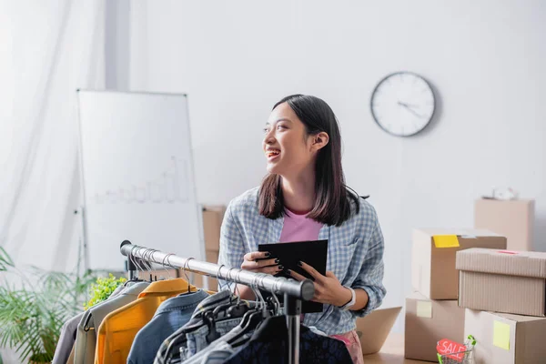 Smiling asian volunteer holding pen and clipboard near clothes and carton boxes on blurred background — Stock Photo