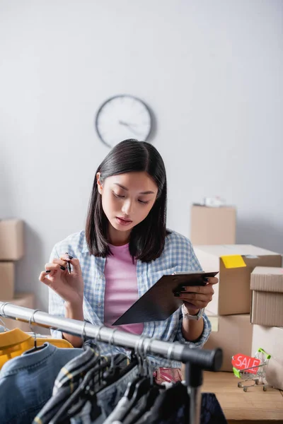 Asian volunteer with clipboard looking at clothes on hanging rack near toy shopping cart with sale on price tag — Stock Photo