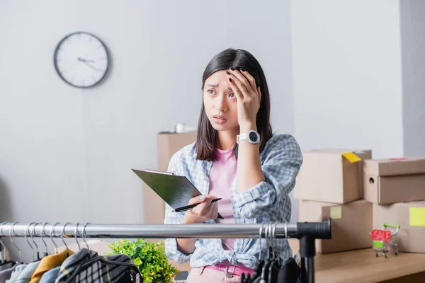 Upset asian volunteer holding clipboard with pen near hanging rack with clothes on blurred foreground in charity center — Stock Photo