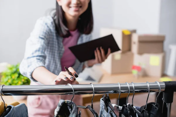 Vista ritagliata del manager che tiene penna e appunti vicino ai vestiti sul rack appeso nel centro di beneficenza su sfondo sfocato — Foto stock