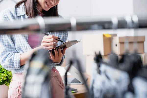 Ausgeschnittene Ansicht eines lächelnden Freiwilligen mit Stift und Klemmbrett, der in der Nähe von Hängegestell auf verschwommenem Vordergrund steht — Stockfoto