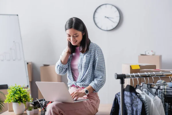 Sorrindo asiático voluntário usando laptop enquanto sentado perto de roupas no pendurado rack no escritório — Fotografia de Stock