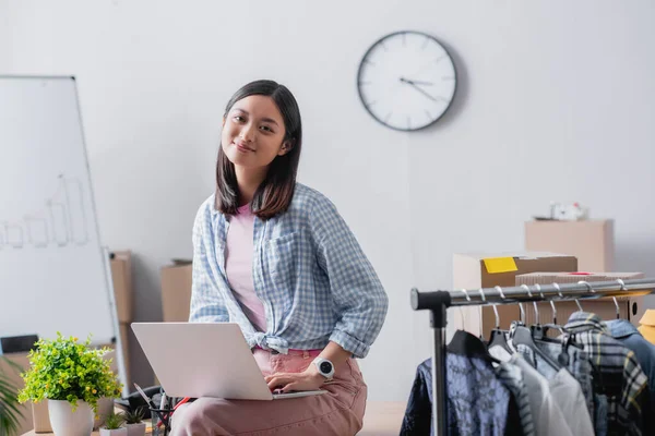 Positivo asiático gerente usando laptop perto de roupas no pendurado rack em primeiro plano desfocado no escritório de caridade — Fotografia de Stock